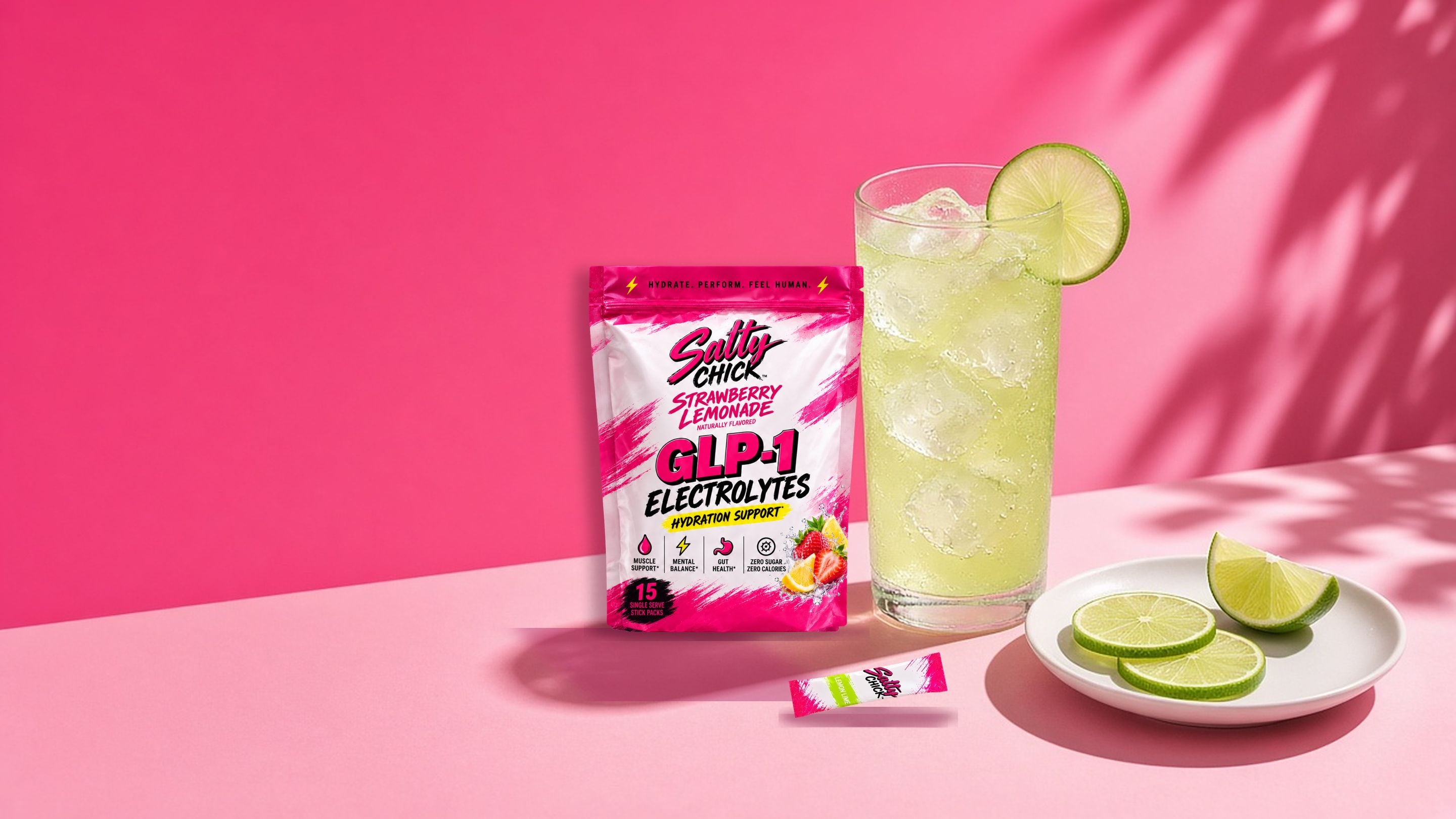 Salty Chick Strawberry Lemonade electrolyte pouch and a Lemon Lime stick pack beside a glass of lemon-lime drink over ice and a plate of lime slices on a pink surface